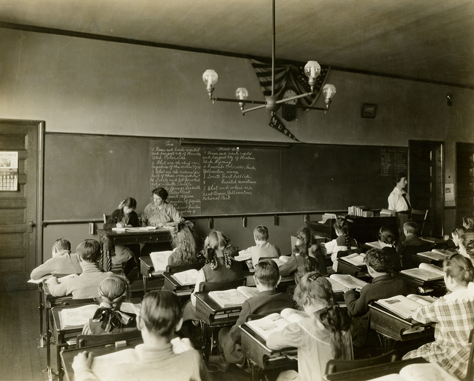 A black and white photograph that features a teacher standing in front of a classroom of students sitting at wooden desks.